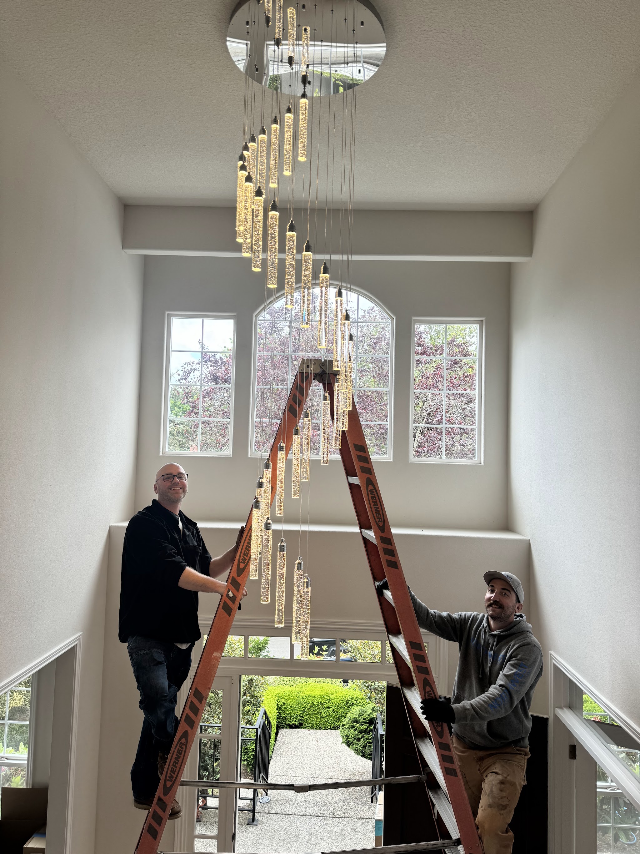 Two technicians installing a large multi-pendant chandelier from a ladder